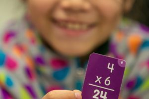 A young child smiling and holding an index card that says, "4 times 6 equals 24."