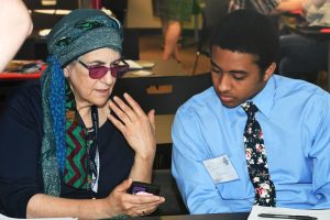 A white teacher with a head scarf showing a black student using an assistive technology device