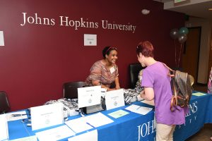 A smiling hispanic woman standing under the "Johns Hopkins University" inscribed on the wall behind an information table inviting a white woman to read about the center