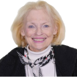 Older white woman with white shoulder length hair wearing a white turtleneck, black blazer, and black and white patterned ascot