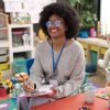 Black female teacher in late 20s wearing glasses sitting at a small children's table in a classroom with a clipboard and pen.