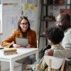 A young white female teacher wearing glasses and holding a clipboard consults with a black father and his tween son at a table.
