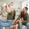 White mom holding up an infant with hispanic father and toddler daughter sitting next to them on a beige couch in a beige family room