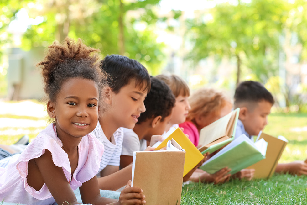 Five children outside in the grass reading books.