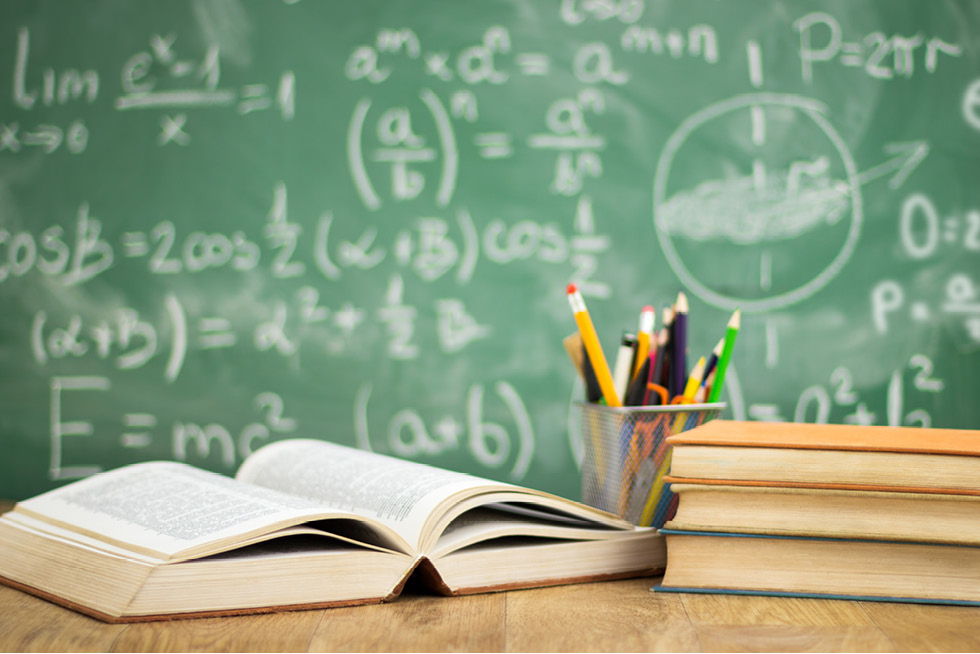 Several books and a cup of pencils on a desk in front of a chalkboard.