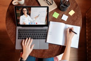 Distance Education Concept. Above top view of female student sitting at desk, writing in notebook, taking notes, using laptop, having online class with teacher.