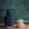 A child and a teddy bear sitting and facing a chalkboard.