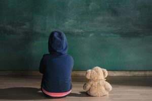 A child and a teddy bear sitting and facing a chalkboard.