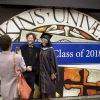 One person taking a photo of two people, one of which is dressed in graduation regalia.