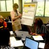 Adults in a room working at a table and on a large notepad.