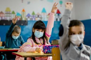 Children wearing masks raising their hands in a classroom.