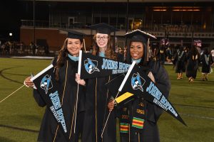 Three people in graduation regalia smiling and holding Johns Hopkins University flags.