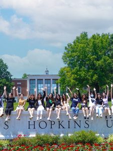 A group of students sitting on the Johns Hopkins University sign on the Homewood campus and raising their hands.