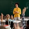 A teacher addressing a classroom while students are raising their hands.