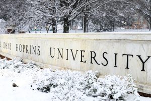Snow covered trees and plants surrounding a marble wall that reads The Johns Hopkins University.