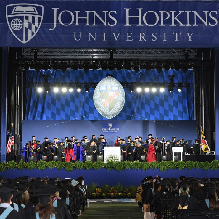 A stage with people sitting in chairs at a graduation with graduates sitting in the crowd.