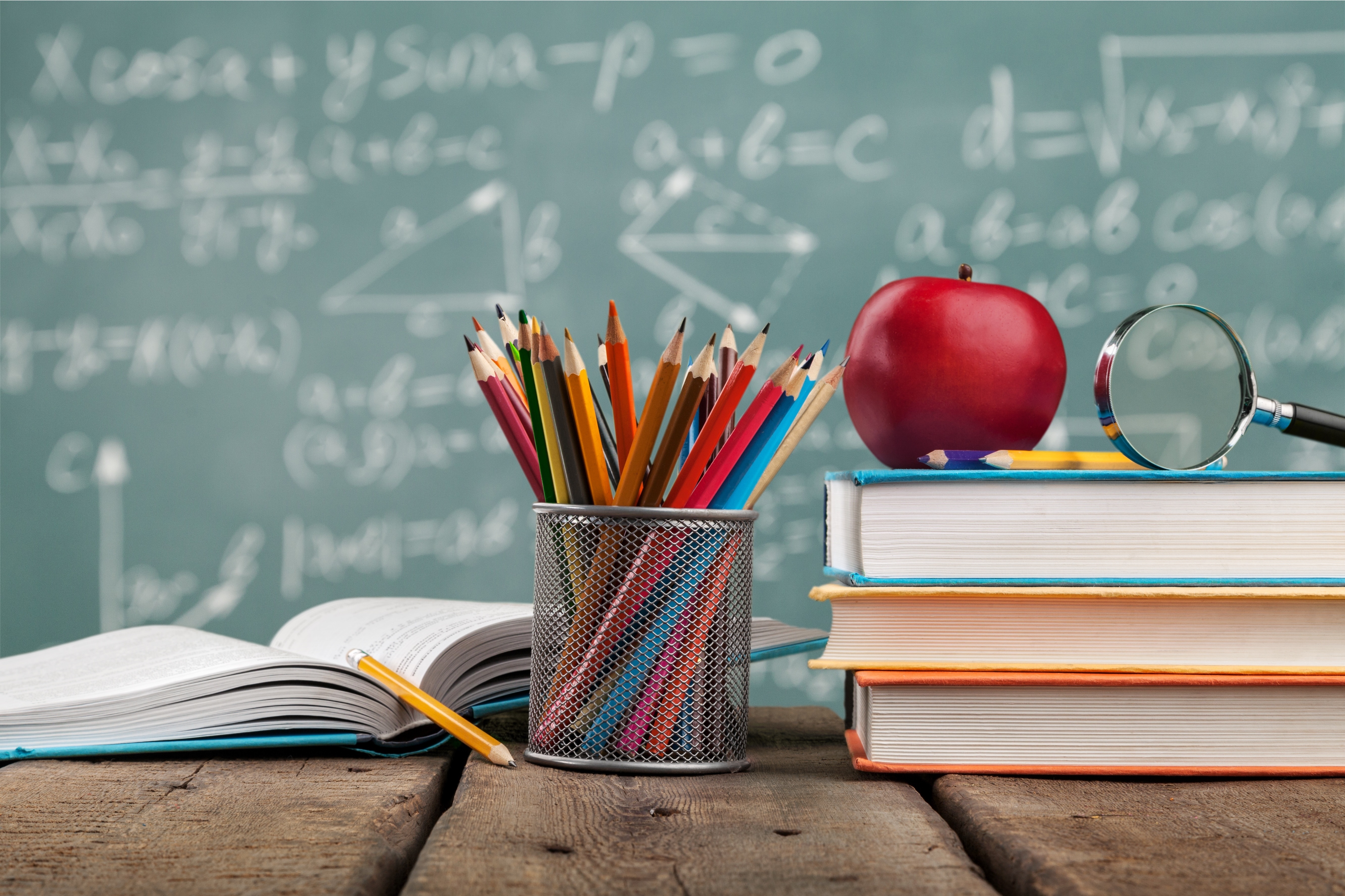 A colorful array of pencils in a mesh cup, an apple, and stacked books on a desk with a blackboard filled with mathematical equations in the background.