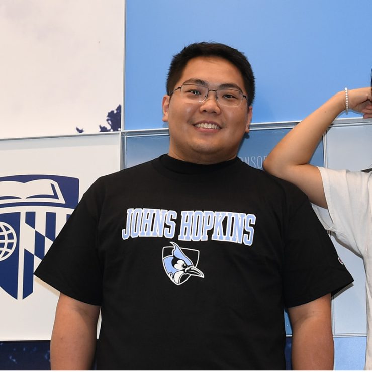 Two people wearing Johns Hopkins t-shirts standing in front of a wall with the Johns Hopkins shield.