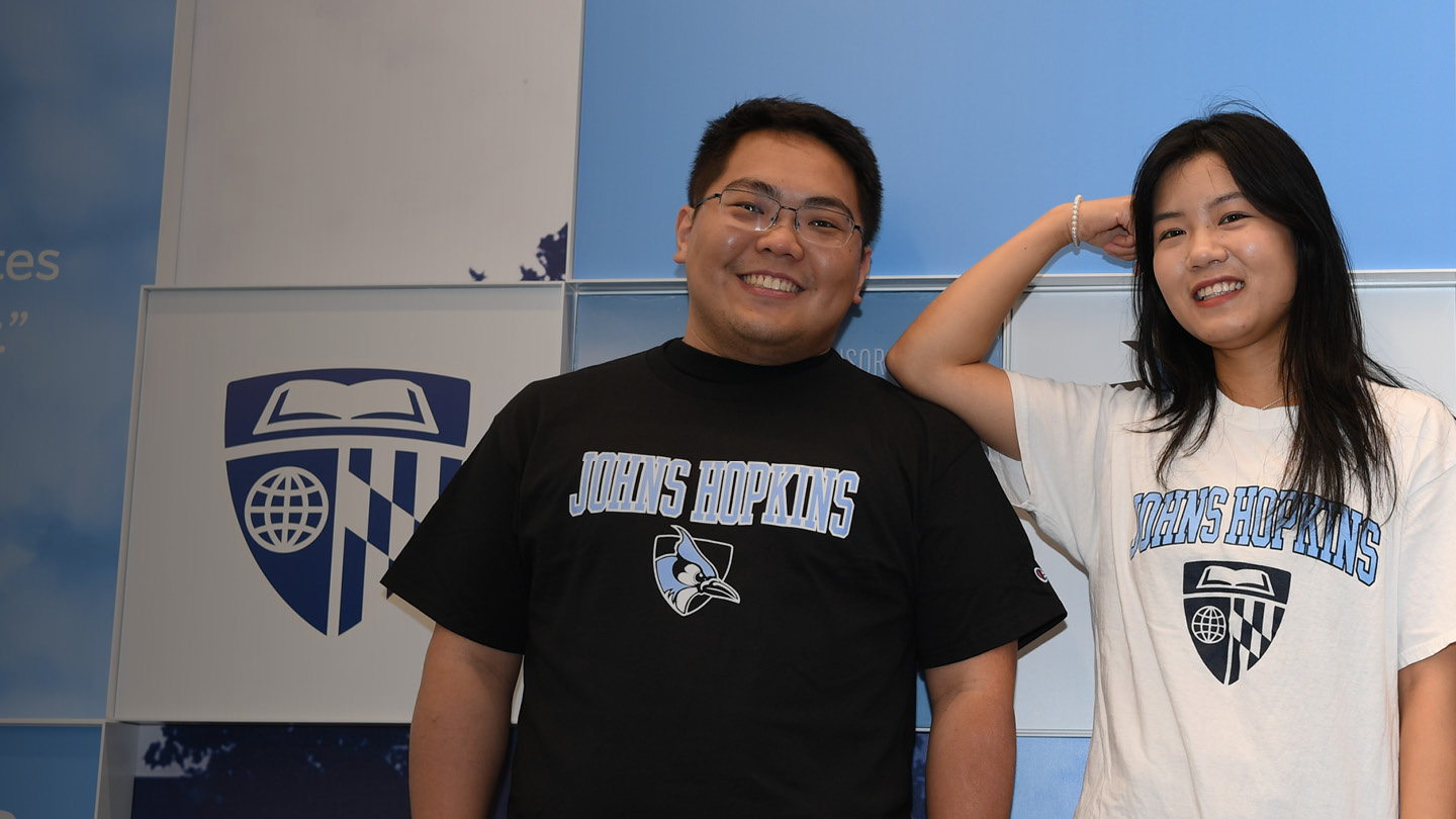 Two people standing in front of a blue wall with the Johns Hopkins shield on it,
