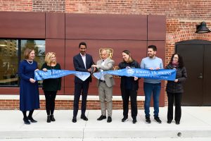 Seven people standing outside of a building holding a large ribbon for the building's reopening