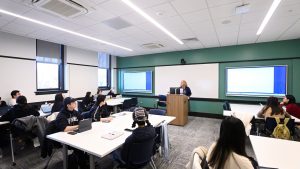 Students sitting at tables in a classroom with an instructor standing at a front podium