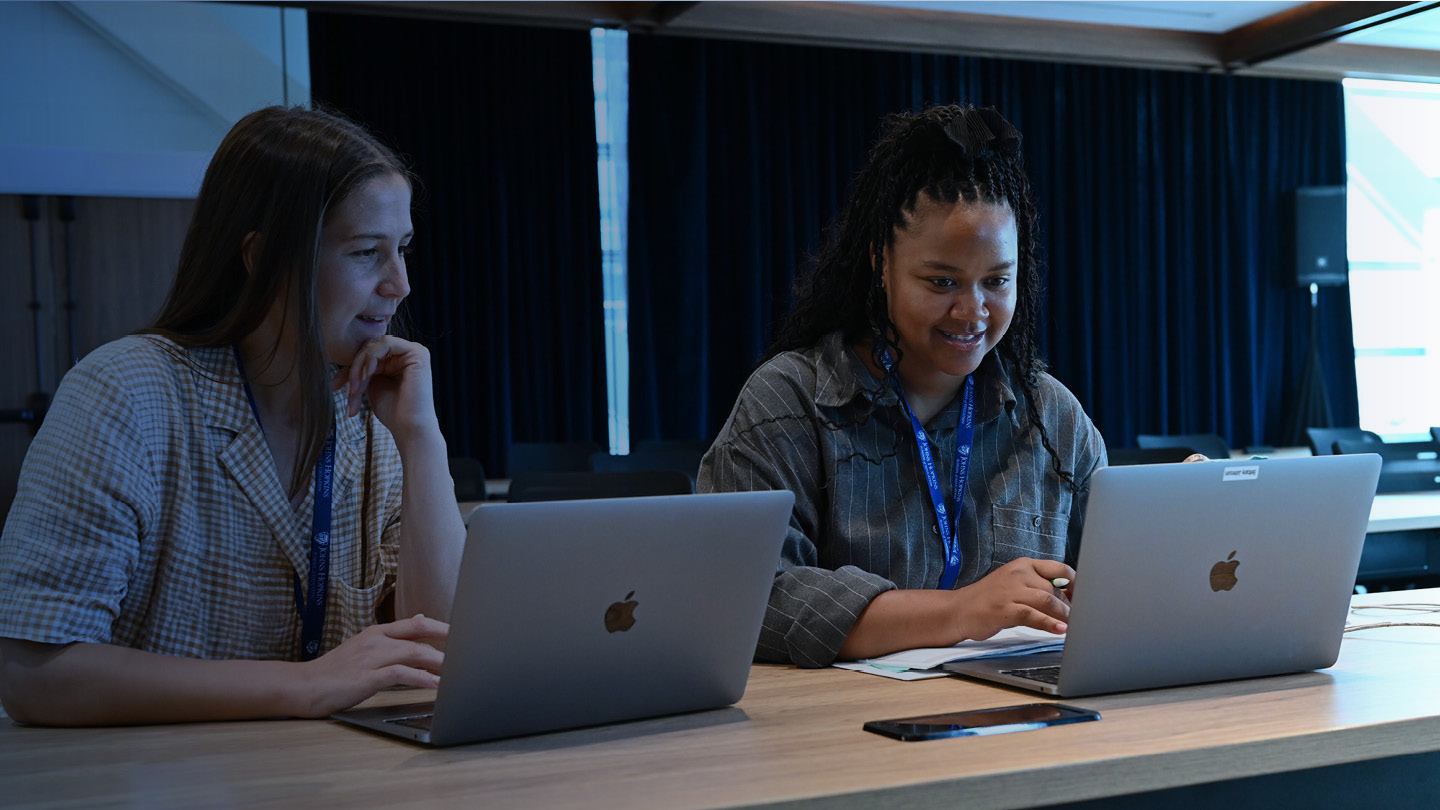 Two people sitting next to each other at a table working on laptop computers